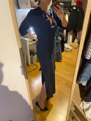 Marinblå långklänning med slits - Marinblå långklänning i linne från Boomerang med knappar ner över bysten. Klänningen har långa ärmar, knytband som kan knytas framtill eller baktill. Slitsar på sidorna till knähöjd. Lantlig stilren look. Storlek 36 men något stor i storleken.