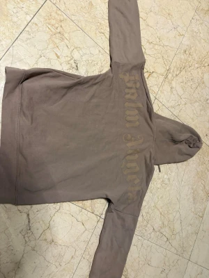 Brun hoodie från Palm Angels - Säljer en brun hoodie från Palm Angels med stor logga i gotisk stil på ryggen. Hoodien har huva med dragsko och en stor magficka framtill. Tillverkad i mjukt bomullsmaterial och har en avslappnad passform. Perfekt för en streetwear-look.