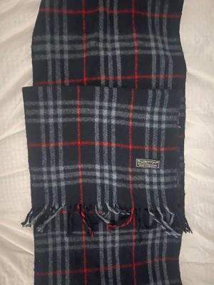 Rutig halsduk från Burberry - Klassisk halsduk från Burberry i mörkblått med rutigt mönster i grått och rött. Halsduken har fransar i ändarna och är tillverkad i mjuk ull. Perfekt accessoar för att lyfta din outfit med en tidlös look. Är använd en gång