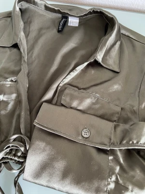 Grön croppad blus - Säljer en croppad blus med glansigt material och knytband i midjan. Blusen har två bröstfickor, krage och långa ärmar med knapp vid ärmslut. Perfekt för dig som vill sticka ut med en trendig look.