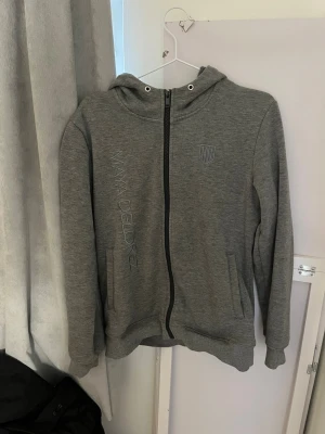 Grå zip-hoodie från Maya Delorez - Grå hoodie med dragkedja från Maya Delorez. Snyggt tryck med logga på bröstet och stort svart X-mönster på ryggen. Två fickor fram och huva med snörning. Tillverkad i mjukt sweatshirtmaterial, perfekt för chill dagar.