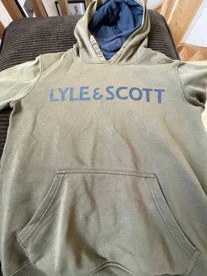 Olivgrön hoodie från Lyle & Scott - Snygg olivgrön hoodie från Lyle & Scott med stor logga i blått över bröstet. Hoodien har en klassisk känguruficka framtill och en fodrad huva i mörkblått tyg. Perfekt för dig som gillar streetwear och vill ha något bekvämt och stilrent.
