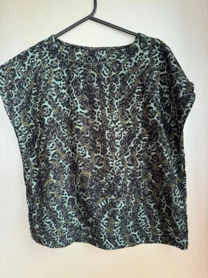Leopardmönstrad topp i grönt och svart - Snygg topp med leopardmönster i gröna och svarta toner. Modellen är lös med rund halsringning roch korta ärmar. Perfekt att ha med sig på resan; strykfri,  tar ingen plats och lätt material.