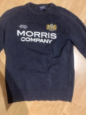 Mörkblå stickad tröja Morris - Mörkblå stickad tröja från Morris med vit text 'Morris Company' och ett broderat emblem i guld och vitt på bröstet. Tröjan har rund halsringning, ribbade muddar och är långärmad. Perfekt för en stilren och klassisk look.