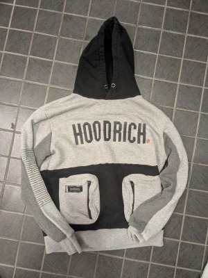 Grå och svart hoodie från Hoodrich - Snygg hoodie från Hoodrich i grått och svart med stor logga på bröstet och textdetaljer på ryggen och ena ärmen. Hoodien har stor huva med dragsko och två framfickor med patch. Perfekt för en streetwear-look.