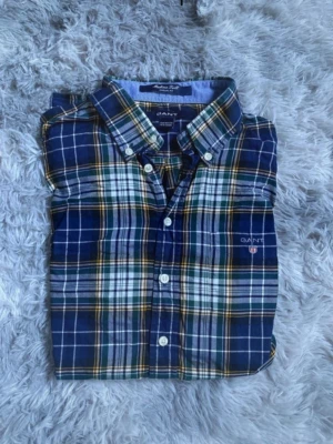 Blårutig skjorta från GANT - Klassisk blårutig skjorta från GANT med vit, gul och grön detalj. Skjortan har knappar framtill, button-down krage och lång ärm. Tillverkad i mjuk bomull och har broderad logga på bröstet. Perfekt för en avslappnad men ändå stilsäker look.priset kan diskuteras vid snabb affär 