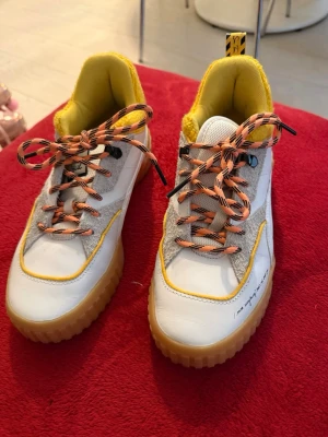 Puma sneakers i vitt och gult - Snygga sneakers från Puma i vitt och gult med orange och svarta snören. Skorna har en robust gummisula, detaljer i mocka och textil samt en mjuk, gul insida. Coola detaljer på hälen och signatur på sidan. Perfekta för dig som vill sticka ut.
