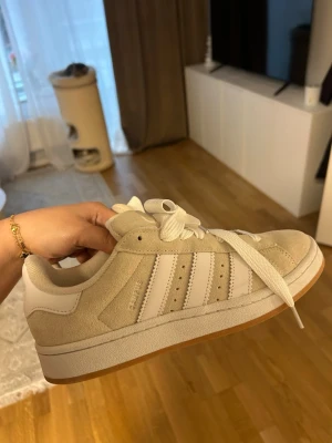 Beige Adidas Campus sneakers - Snygga Adidas Campus sneakers i beige mocka med vita detaljer och klassiska tre ränder på sidan. Skorna har vita skosnören, gummisula och låg profil. Perfekta för en clean och stilren look. Adidas-logga på plösen och insidan.