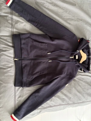 Marinblå hoodie från Moncler - Snygg marinblå hoodie från Moncler med dragkedja, huva med dragsko och två fickor framtill. Ärmsluten har vita och röda detaljer och Moncler-loggan sitter på vänster ärm. Tillverkad i mjuk bomull för skön känsla.