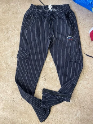 Svarta cargobyxor med vita ränder - Säljer ett par svarta cargobyxor med vita tunna vertikala ränder. Byxorna har elastisk midja med dragsko, sidofickor med dragkedja och klassiska cargofickor på benen. Detalj med broderad patch på ena benet och dragkedja vid benslut. Perfekta för en avslappnad streetstil.