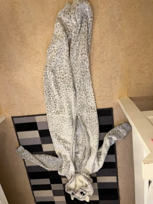 Grå fläckig djuronesie med huva - Mysig grå onesie i plysch med svarta och vita fläckar, dragkedja framtill och huva med söta djuröron och broderade ögon. Perfekt för maskerad eller cosplay när du vill vara ett gulligt djur.