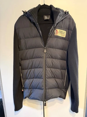 Moncler Grenoble Cardigan  - Svart cardigan från Moncler Grenoble med vadderad front, stickade ärmar och huva. Jackan har dragkedja, patch med Moncler-logga på bröstet och ärmen samt ribbad krage. Materialmix av dun och stickat ger en exklusiv och sportig känsla. Inköpt på NK Stockholm, sparsamt använd.