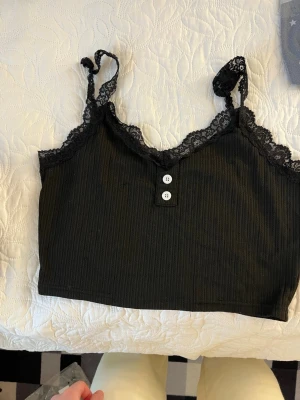 Svart ribbad linnetopp med spets - Trendig svart ribbad linnetopp med spetskant och smala axelband. Toppen har två dekorativa knappar framtill och är croppad för en modern look. Perfekt att styla med höga jeans eller kjol.