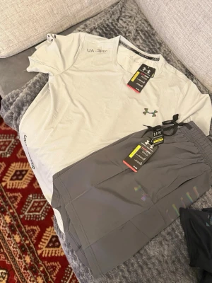 Under Armour träningsset t-shirt & shorts - Snyggt träningsset från Under Armour med en ljusgrå t-shirt och mörkgrå shorts. T-shirten har UA-Spec-tryck och logga på bröstet, medan shortsen har elastisk midja med snörning och diskret logga. Perfekt för gymmet eller löpning.