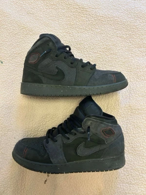 Nike Air Jordan 1 Mid mörkgrå - Snygga Nike Air Jordan 1 Mid sneakers i mörkgrå mocka och läder. Skorna har svarta detaljer, diskreta röda sömmar vid tån och hälen samt klassisk Nike Swoosh på sidan. Perfekta för dig som gillar streetwear och vill ha en stilren look. Köpte den från amerikanska JD för 1600kr