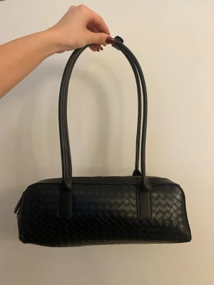 Svart axelväska med flätad design - Snygg svart handväska med lång axelrem och flätad läderlook. Väskan har rektangulär form, guldfärgad dragkedja och rymligt innerfack. Perfekt för dig som gillar stilrena accessoarer med en twist.