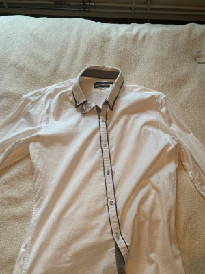 Vit skjorta från Celio Club M/L - Snygg vit skjorta från Celio Club i stretchigt material. Skjortan har lång ärm, svarta detaljer längs knappraden och kragen samt diskret rutigt mönster på insidan av kragen. Perfekt för en clean och stilren look.