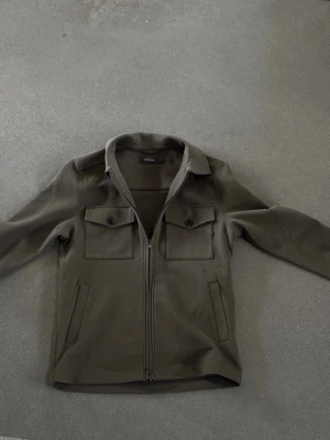 Olivgrön overshirt med dragkedja - Stilren olivgrön overshirt från dressman med två bröstfickor med lock och knapp, samt dragkedja framtill. Klassisk krage och raka ärmar. Perfekt lager på lager-plagg i mjukt material med minimalistisk look.