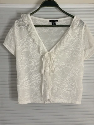Vit spetsig bolerotopp från Amisu - Säljer en vit bolerotopp från Amisu i genombruten spets med korta ärmar och v-ringning. Toppen har volangkant och knyt framtill för en söt och romantisk look. Perfekt att styla över ett linne eller klänning.