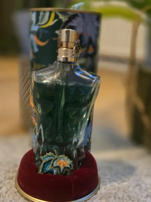 Jean Paul Gaultier Le Male EdT - Unik parfymflaska från Jean Paul Gaultier Le Male, formad som en manlig torso i genomskinligt glas med grönblå vätska. Flaskan har broderade detaljer och kommer med en dekorativ tub med blommigt mönster. Perfekt för dig som gillar ikoniska dofter och snygg design.