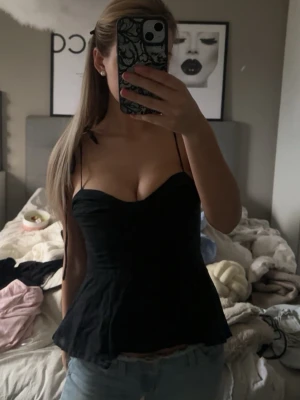 Svart korsettopp med tunna band - Snygg svart korsettopp med tunna axelband och figurnära passform. Toppen har en lätt utsvängd nederdel och markerad byst, vilket ger en feminin siluett. Perfekt att styla med jeans eller kjol för en trendig look.