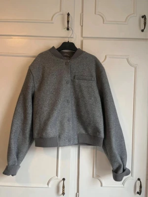 Grå cardiganjacka med tryckknappar - Snygg grå cardiganjacka med ribbade muddar och krage. Jackan har tryckknappar framtill och en bröstficka för en clean look. Materialet är mjukt och tjockt, perfekt för lager på lager. Passar dig som gillar minimalistisk och stilren design.