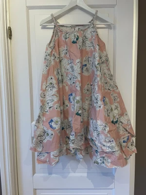 Blommig rosa sommarklänning - Luftig och svingig rosa klänning med tunna axelband och blommigt mönster i vitt, blått och grönt. Klänningen har en vid passform och volangkant nedtill, perfekt för varma dagar. Storlek är 158/164. 