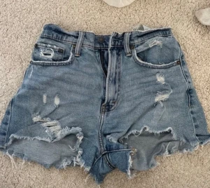 Blå jeansshorts Abercrombie & Fitch - Säljer ett par blå high rise jeansshorts från Abercrombie & Fitch, modell The Mom Short Curve Love. Shortsen har slitna detaljer, råa kanter och fem fickor. Perfekta för en avslappnad och trendig sommarstil.