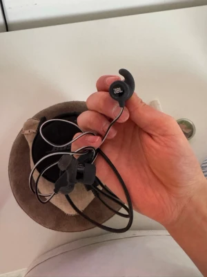 JBL svarta sporthörlurar in-ear - Säljer ett par svarta in-ear hörlurar från JBL med sportig design och öronkrokar för extra bra passform. Sladden är tunn och flexibel, och hörlurarna kommer med ett smidigtu svart fodral för enkel förvaring. Perfekta för träning eller vardagslyssning.