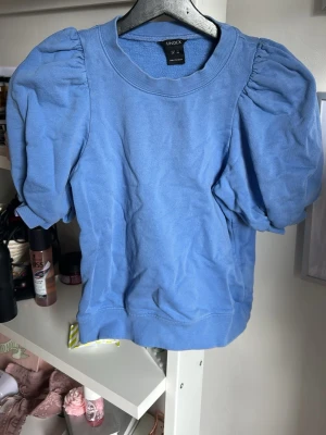 Blå puffärmad topp från Lindex - Snygg blå topp från Lindex med puffiga korta ärmar och rund halsringning. Tillverkad i mjukt material som känns skönt mot huden. Perfekt för dig som vill ha en trendig och bekväm look.