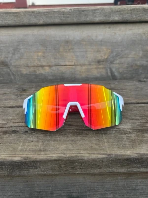 Röd gul blå sportiga solglasögon unisex  - Säljer ett par riktigt färgstarka och sportiga solglasögon med spegelglas i skiftande färger och vit båge. Glasögonen har en futuristisk, kantig form och passar perfekt för dig som vill sticka ut. Snygg accessoar till sommaren! Nya, unisex. One size för vuxna