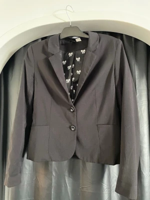 Svart kavaj med rosett-mönstrat foder - Stilren svart kavaj med två knappar framtill och klassisk krage. Insidan har ett svart foder med vita rosetter som ger en lekfull touch. Kavajen har två fickor och en figurnära passform, perfekt för en trendig och snygg look.