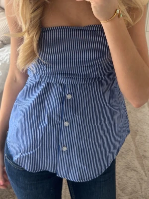 Blåvit randig bandeau tubtopp - Säljer en blå och vit randig bandeau tubtopp med knappar framtill från new Yorker. Toppen är ärmlös och har en avslappnad passform, perfekt för sommardagar. Snygg till både jeans och kjol! Knappt använd då den är för liten!