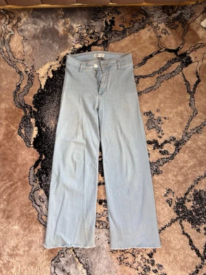 Ljusblå raka jeans med rå kant - Säljer ett par ljusblå jeans med rak passform och rå, fransad kant nertill. Jeansen har klassisk femficksdesign och stängs med knapp och dragkedja. Perfekta för en avslappnad och trendig look. Funkar perfekt för en dag på stranden. Anledningen till att jag säljer är på grund av att den är för stor på mig. 