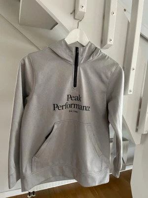 Grå hoodie från Peak Performance - Snygg grå hoodie från Peak Performance med svart logga på bröstet och halv dragkedja framtill. Hoodien har stor känguruficka och huva. Tillverkad i mjukt material som känns skönt mot huden. Perfekt för en avslappnad och sportig stil.