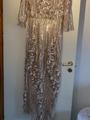 Festklänning med paljetter - Säljer en helt ny elegant festklänning i champagnefärg med glittriga paljetter och blommigt mönster. Klänningen har trekvartsärm och är täckt av skimrande detaljer som verkligen sticker ut. Perfekt för speciella tillfällen där du vill glänsa.