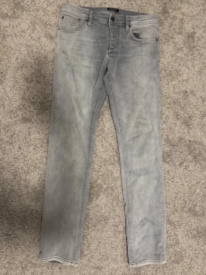 jeans  Jack & Jones - Säljer ett par grå slim fit jeans från Jack & Jones. Jeansen har klassisk femficksmodell, normal midja och dragkedjegylf. Tillverkade i mjuk denim med lätt tvättad look och raka ben. Midjemått 80 cm och innebenslängd 82cm.