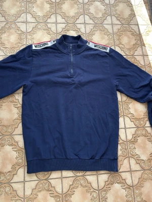 Marinblå half-zip hoodie Moschino - Snygg marinblå hoodie från Moschino med half-zip framtill och ribbad krage. Axelpartiet har vita band med Moschino-logga och en röd detalj längs kanten. Tillverkad i mjukt bomullsmaterial och har långa ärmar samt ribbade muddar.