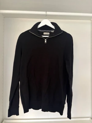 Svart stickad tröja från Jack & Jones - Snygg svart stickad tröja från Jack & Jones med halv dragkedja vid halsen och hög krage. Tröjan har lång ärm och ribbade muddar vid ärmslut och nederkant. Perfekt till lager på lager och enkel att matcha med olika outfits.