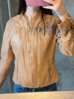 Ljusbrun skinnjacka med dragkedjor - Snygg ljusbrun skinnjacka med fyra dragkedjefickor framtill och ståkrage. Jackan har figurnära passform och detaljerade sömmar som ger en modern look. Perfekt till jeans och enkel att matcha med olika stilar. Passar till mig som bär storlek S/XS.