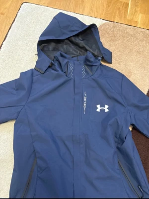 Blå vindjacka från Under Armour - Snygg blå vindjacka från Under Armour med huva och dragkedja. Jackan är helt ny och inga skador alls(finns tagg med) OBS (på jackan står det storlek M men det är storlek S)
