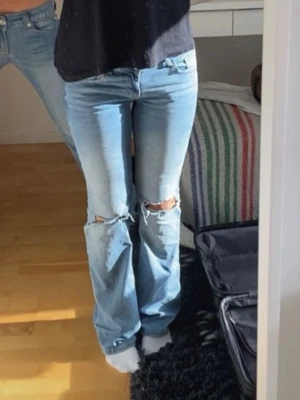 Ljusblå slitna bootcut jeans - Säljer ett par ljusblå bootcut jeans med slitningar och hål vid knäna. Jeansen har klassisk femficksdesign och är tillverkade i denim med en avslappnad passform. Perfekta för en trendig och avslappnad look. Fickorna på rumpan har överlappsfickor med knapp , de är Lågmidjade 