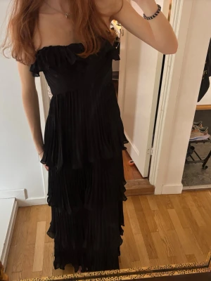 Svart offshoulder långklänning - Säljer en svart långklänning med offshoulder-design och volangdetaljer upptill och längs kjolen. Klänningen har flera lager och ett plisserat tyg som ger en elegant look. Går att ha utan band som bandeau (första bilden) eller med band som på de andra bilderna! Varan är i aldrig använd och har lappen kvar!