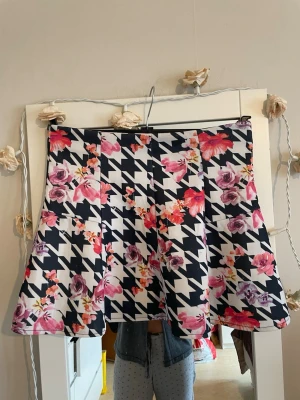 FB Sister svart, vit, rosa, lila kjol, storlek M - Säljer en kort kjol med svartvitt hundtandsmönster och färgglada blommor i rosa, lila och rött. Kjolen har en lätt utställd form och är tillverkad i ett mjukt, följsamt material. Perfekt för dig som vill sticka ut med en unik och trendig look.