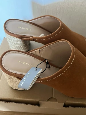 Camel slip-in sandaler från Parfois 7cm - Snygga bruna slip-in sandaler från Parfois med öppen tå och bred rem i mockaimitation. Sandalerna har en chunky klack i flätad jute-look och kontrastsömmar längs kanten. Perfekta för en trendig och avslappnad stil.