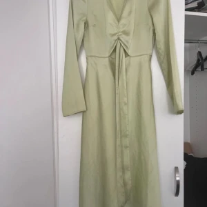 Ljusgrön långklänning med knyt - Säljer en ljusgrön långklänning i satinliknande material med långa ärmar och snygg v-ringning. Klänningen har knyt framtill och en lätt glansig finish som ger en elegant look. 