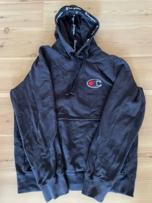 Svart Champion hoodie med logga - Snygg svart hoodie från Champion med stor broderad logga på bröstet och mindre logga på ärmen. Huva med dragsko och Champion-tryck på insidan. Klassisk känguruficka framtill och ribbade muddar. Perfekt för en chill och sportig look. Det är large för barn. 