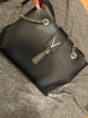 Valentino handväska läder svart kvinna - Valentino handväska i svart läder för kvinna. Väskan har kedjehandtag i metall och dekorativ tassel framtill. Invändigt finns flera fack, inklusive ett större huvudfack, ett innerfack med dragkedja samt mindre fack för exempelvis mobil och småsaker. Passar för vardagsbruk och tillfällen där det behövs plats för personliga tillhörigheter som plånbok, nycklar och mindre accessoarer.