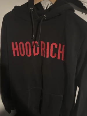 Svart HOODRICH hoodie med röd text - Snygg svart hoodie från Hoodrich med stor röd logga på bröstet. Klassisk huva med dragsko och magficka framtill. Perfekt för en streetwear-look och riktigt skön att bära till jeans eller joggers. Kan skickas även mötas upp i Stockholm