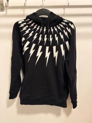Neil Barrett Hoodie - Cool svart hoodie med vita blixtar som strålar ut från halsen både fram och bak. Tröjan har huva med snören och ribbade muddar vid ärmslut och nederkant. Perfekt för dig som vill sticka ut med en edgy look.